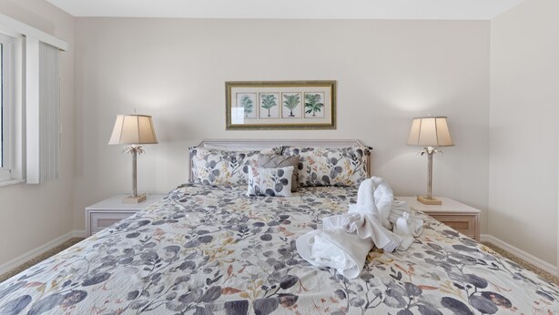 A bedroom with a bed, two bedside lamps, a framed botanical print above the bed, and a window with blinds. - Sand Caper #702 (Fort Myers Beach)