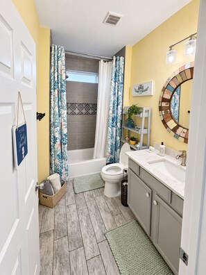 A bathroom with a bathtub, toilet, and sink, featuring a wooden floor and patterned curtains. - Chasing Ray's 2-bedroom, 2-Bath cottage in Mexico Beach, 3 min walk to beach. (Mexico Beach)