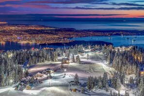 A snow-covered ski resort with a panoramic view of a city at dusk.