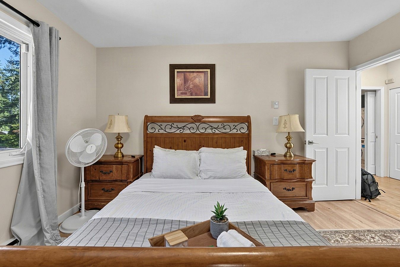 A bedroom with a wooden bed, two bedside tables with lamps, a window with curtains, a door, and a small potted plant on the bed.