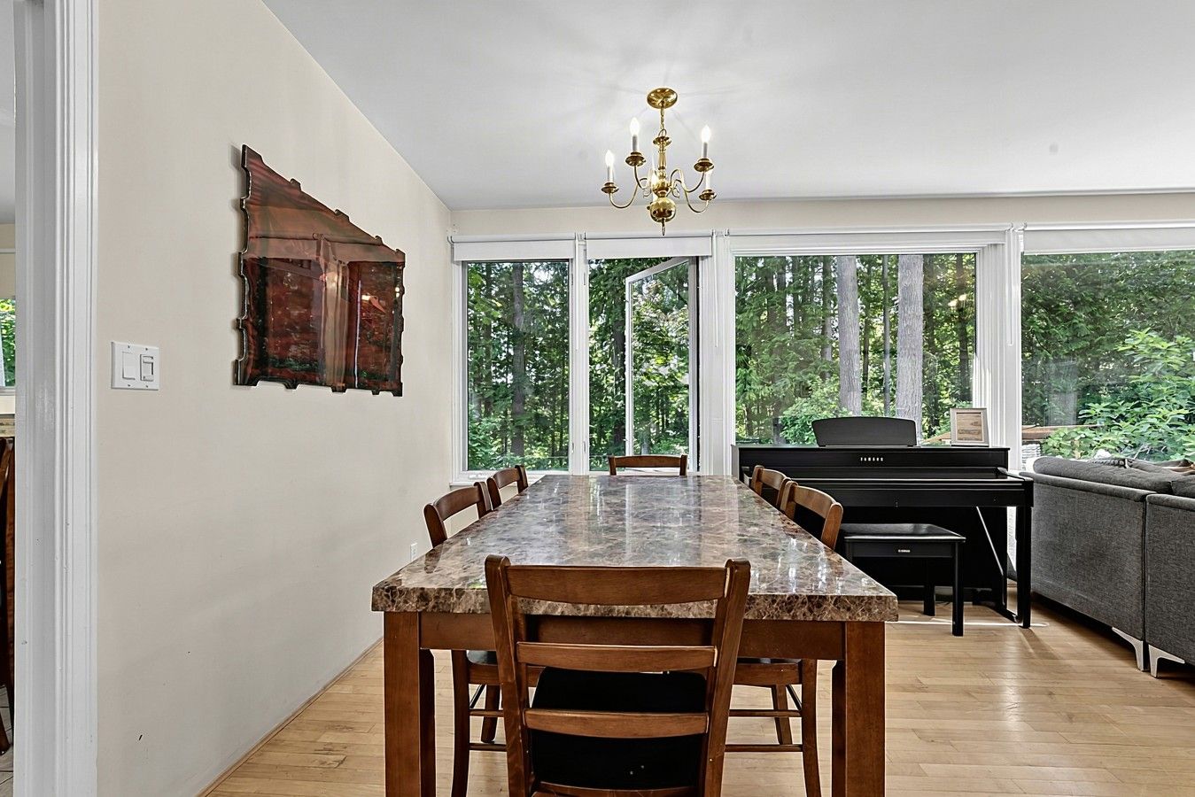 Un espace repas comprenant une table en marbre, des chaises en bois, un piano et une vue sur la forêt grâce à de grandes fenêtres.