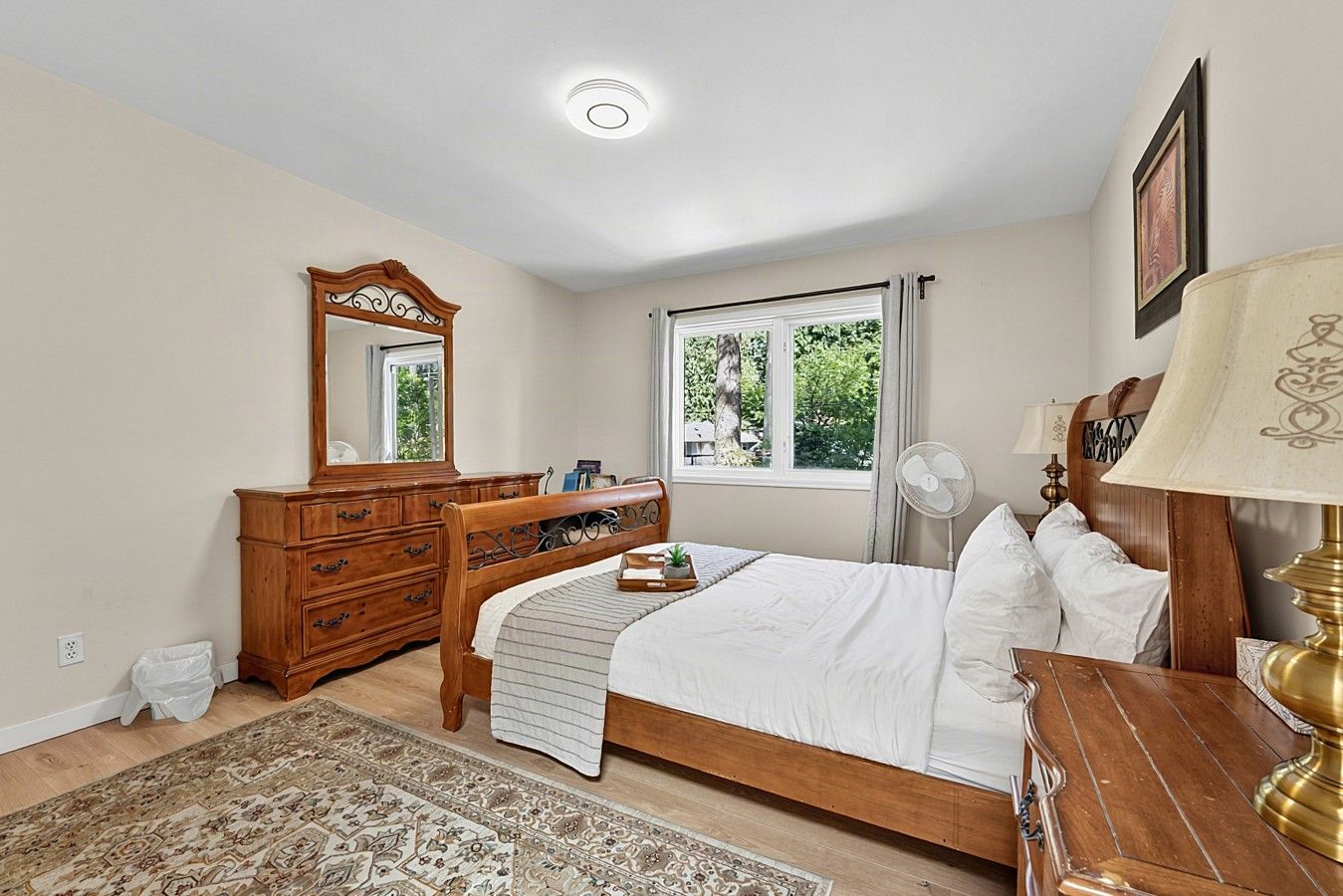 A bedroom with a wooden bed, a dresser, a mirror, a window with curtains, a fan, and a rug.