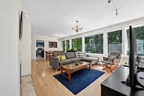 A modern living room with a sectional sofa, a coffee table, and a TV. There is a dining area with a table and chairs, and a kitchen in the background.