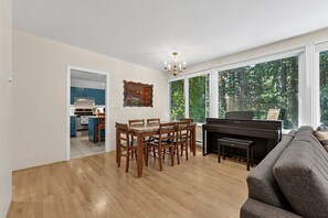 Un salon avec un piano, une table à manger et des chaises, une cuisine à l’arrière-plan et de grandes fenêtres donnant sur la forêt.
