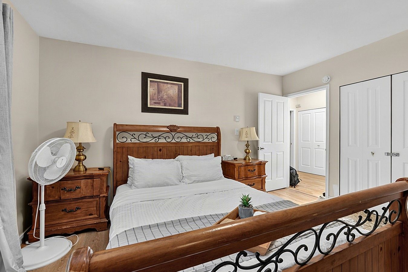 A bedroom with a wooden bed, two bedside tables with lamps, a white fan, a framed picture on the wall, and a view of a hallway with a door.