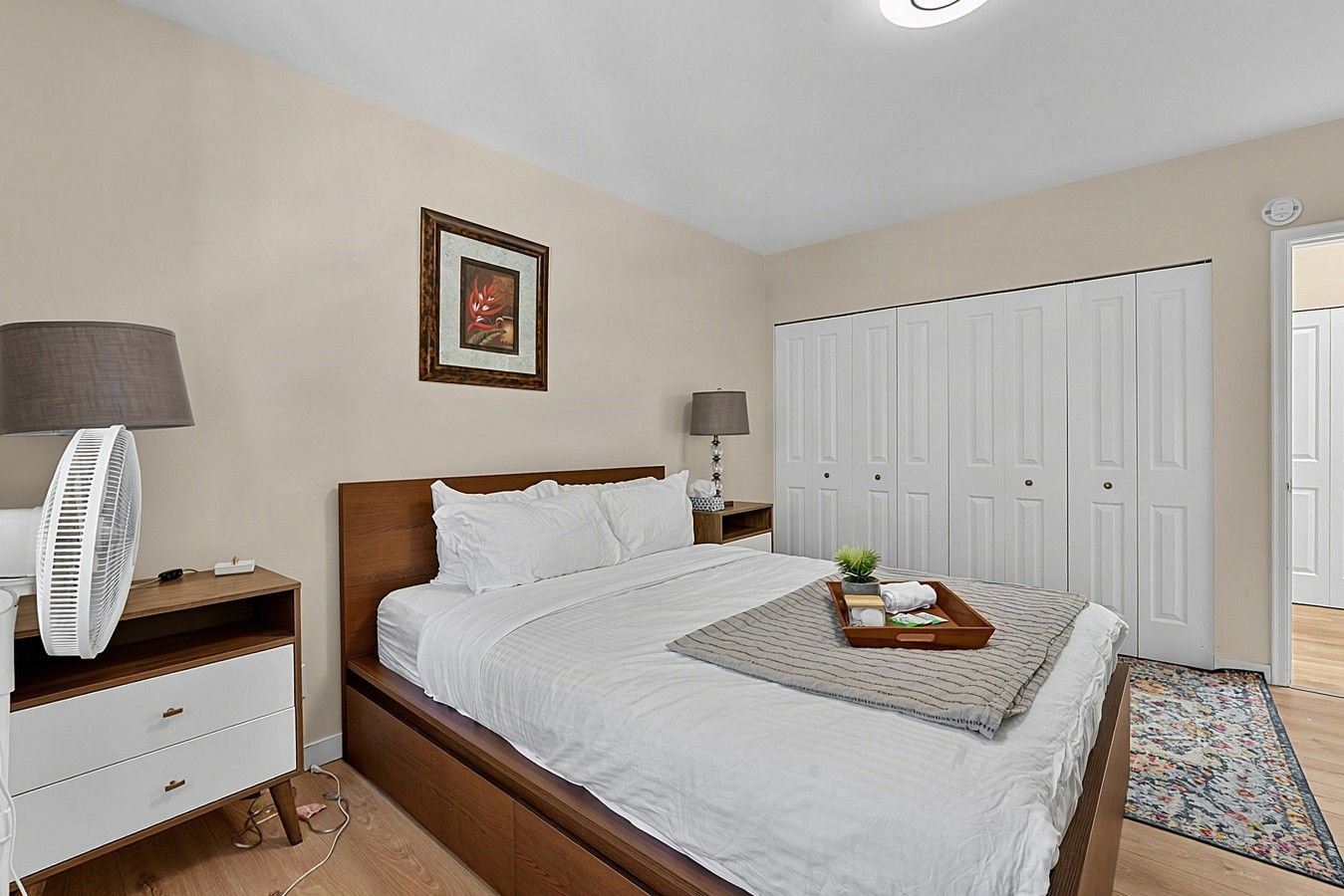 A neatly arranged bedroom with a bed, bedside tables, a fan, a framed picture, a wardrobe, and a small plant on the floor.