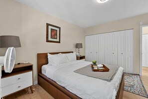 Une chambre bien rangée, avec un lit, des tables de chevet, un ventilateur, un tableau encadré, une armoire et une petite plante posée sur le sol.
