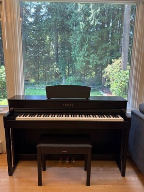 Un piano droit Yamaha avec un banc, dans une pièce dotée d’une grande fenêtre donnant sur une forêt.
