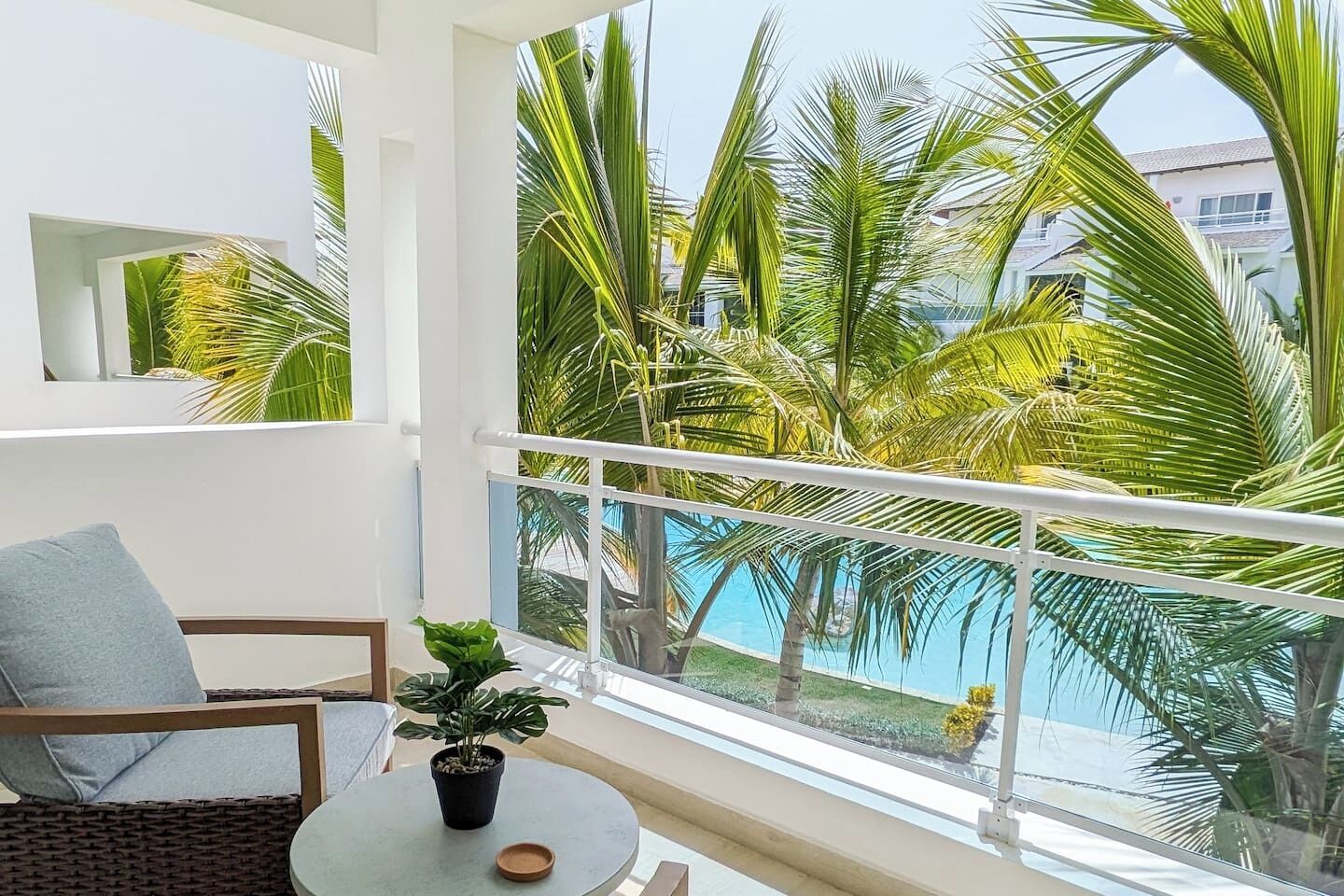 Un balcón con vista a una piscina y palmeras, una mesa redonda con una planta en maceta y un sofá gris.
