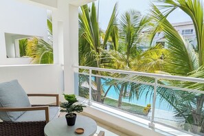 Un balcón con vista a una piscina y palmeras, una mesa redonda con una planta en maceta y un sofá gris.