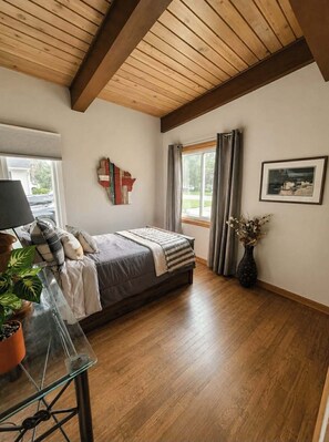 A bedroom with a bed, a window with curtains, a vase with flowers, and a framed picture on the wall.
