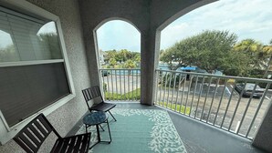A balcony with two chairs, a patterned rug, and a view of a parking lot and trees. - Ocean Gate I: Cozy Retreat Near Beach (St. Augustine)