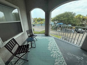 A balcony with two chairs, a patterned rug, and a view of a parking lot and trees. - Ocean Gate I: Cozy Retreat Near Beach (St. Augustine)