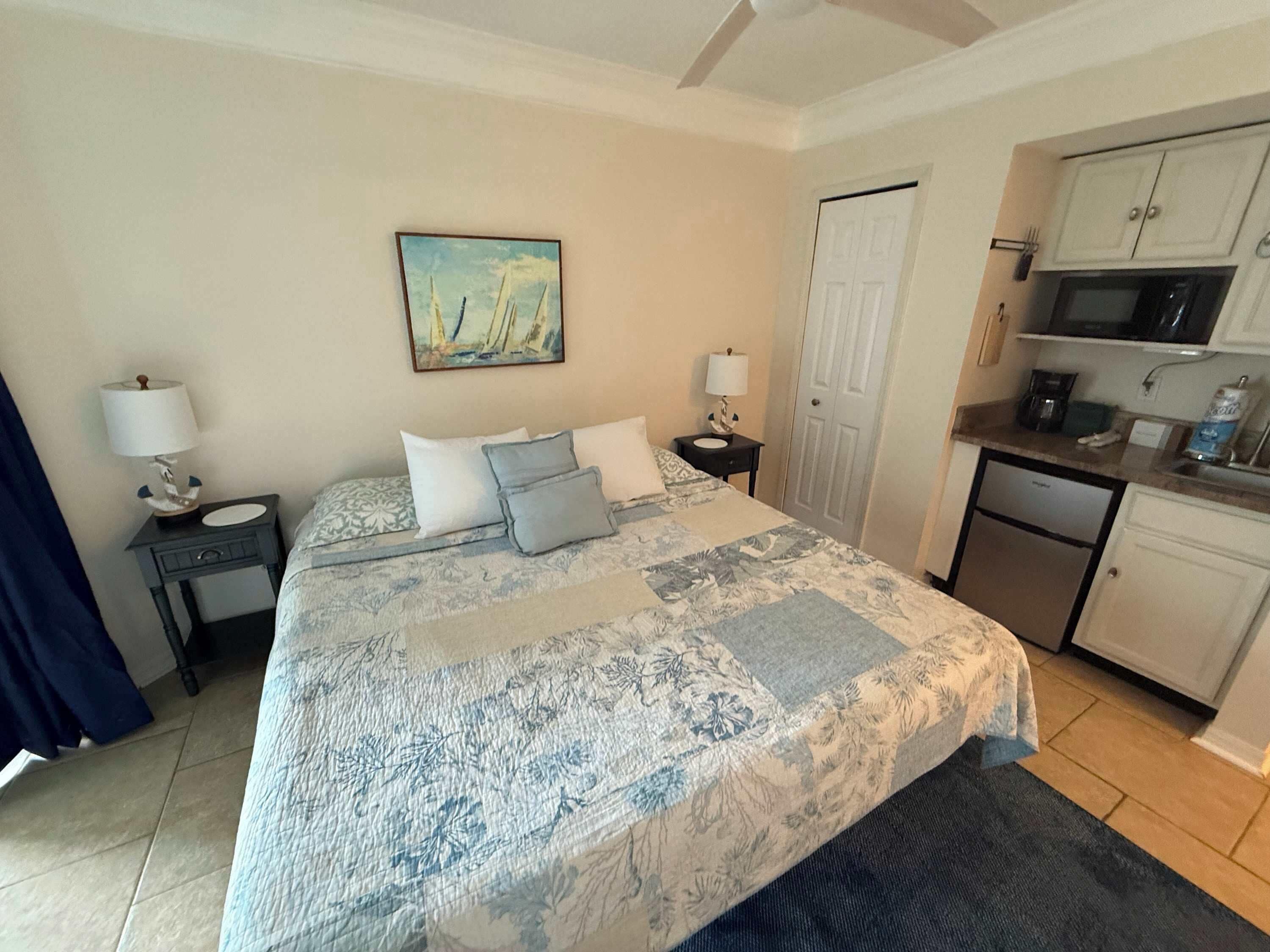 Un dormitorio con cama, mesita de noche, un cuadro, una puerta y una kitchenette.