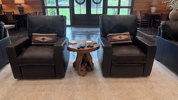 A cozy interior with two leather armchairs facing a wooden coffee table, a large window with a view of greenery, and a door with wreaths.