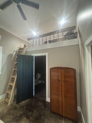 A room with a ceiling fan, a wooden ladder, a wardrobe, and a door leading to a dark space.