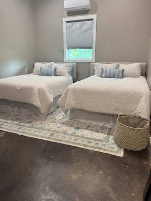 A bedroom with two beds, a window with blinds, a woven basket, and a patterned rug.