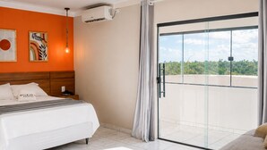 A hotel room with a bed, a nightstand, an air conditioning unit, a window with a view, and a curtain.