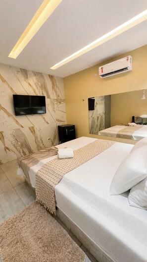 Habitación de hotel con televisor de pantalla plana en la pared, cama con ropa de cama blanca, alfombra color beige y unidad de aire acondicionado.