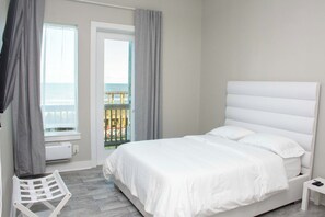 Ein Schlafzimmer mit einem weißen Bett, einem kleinen weißen Nachttisch, einem weißen Stuhl und Meerblick durch eine Balkontür.