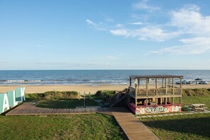 Eine Holzkonstruktion am Strand mit einer Terrasse und Treppe, ein Weg führt dazu, und es gibt einen klaren Blick auf das Meer.