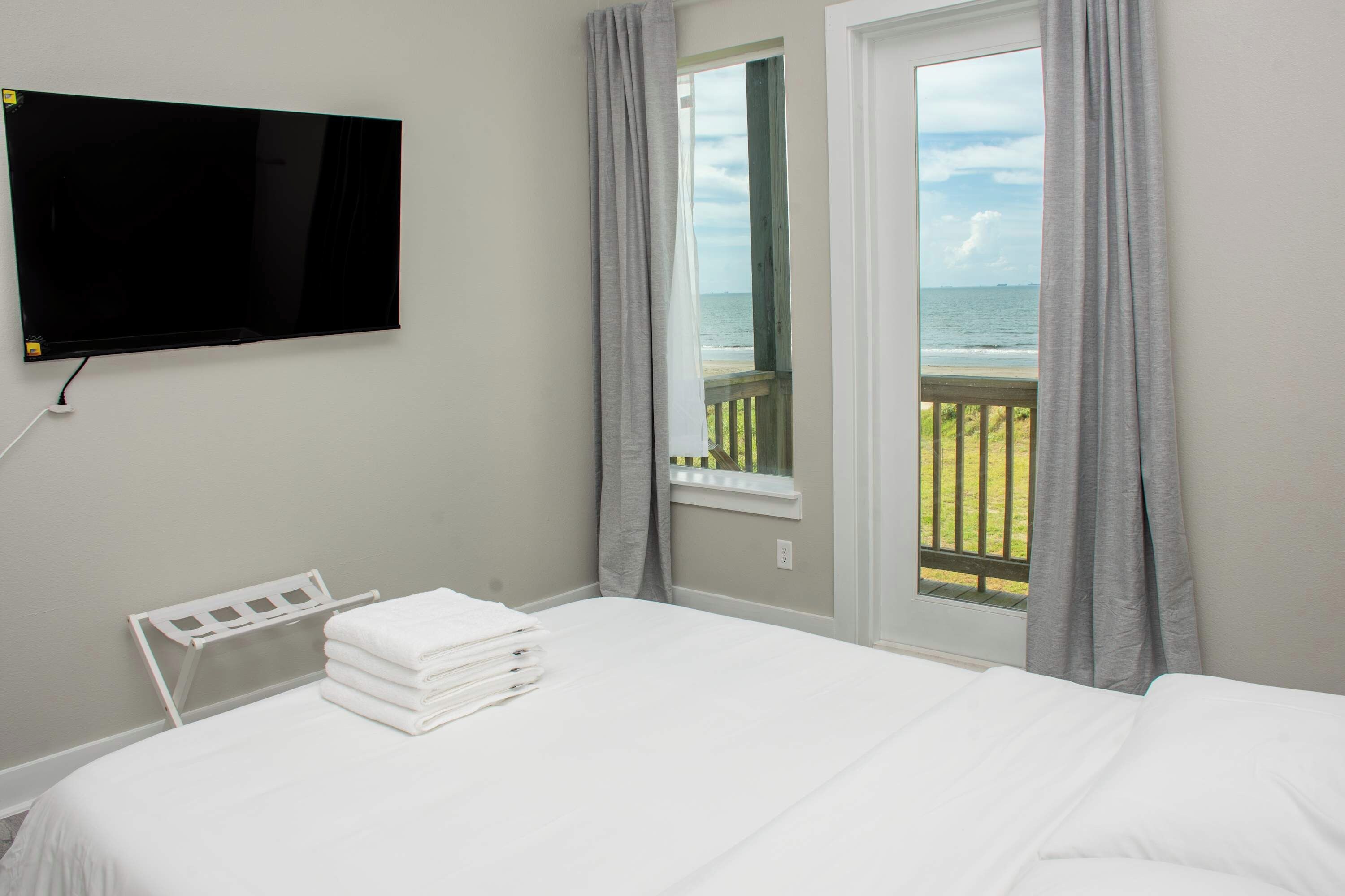 A hotel room with a bed, a TV mounted on the wall, a window with curtains, and a view of the sea.