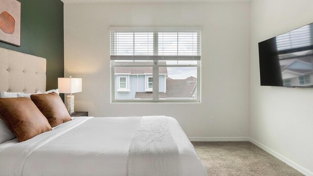 A hotel room with a bed, a nightstand, a lamp, a window with blinds, and a flat-screen TV mounted on the wall. - Landing | Amazing 2BD, Gym, Yoga Studio (Four Corners)