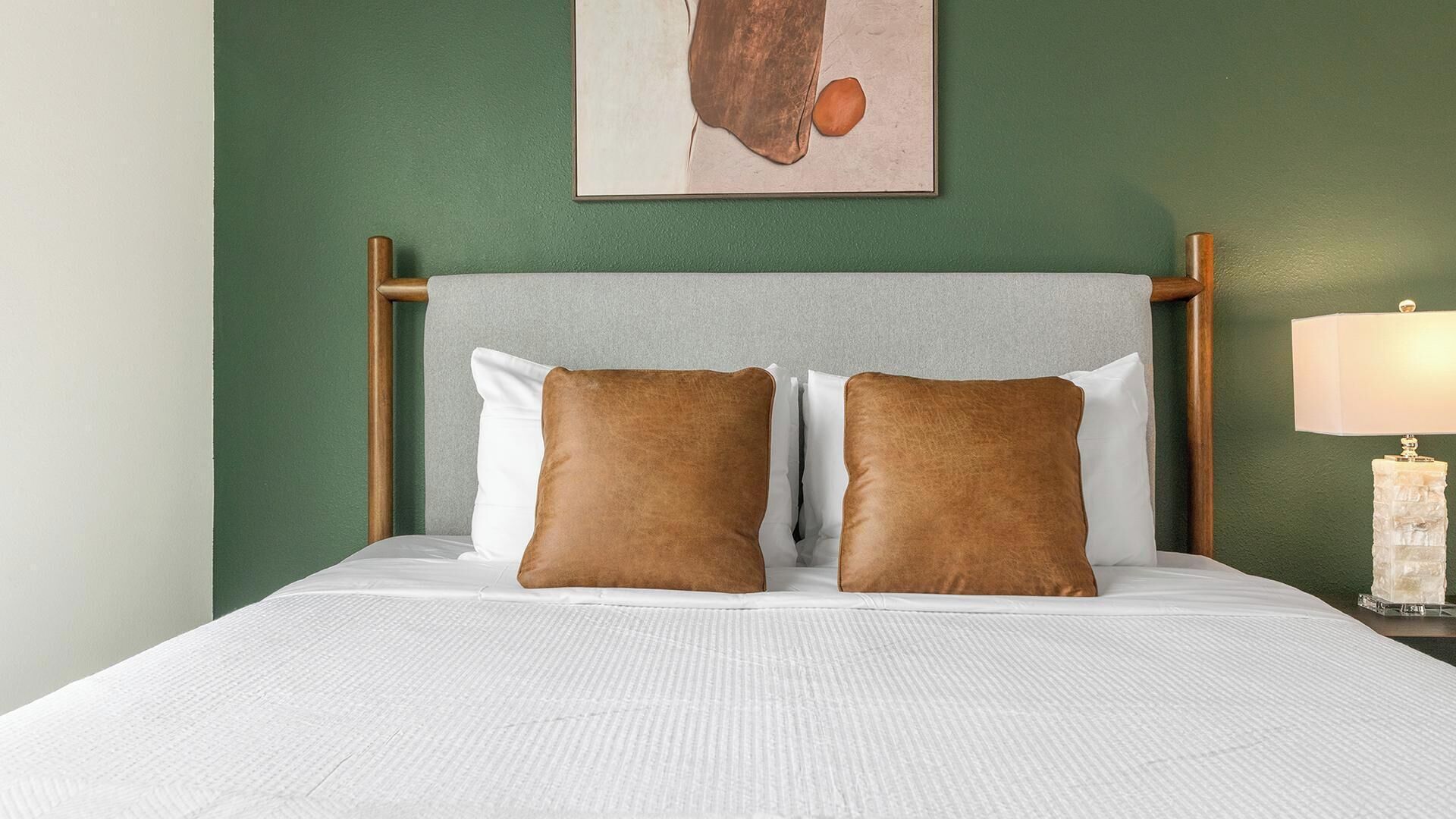 A bed with a grey headboard, two brown pillows, white bedding, and a framed picture on the wall.