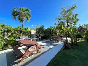 An outdoor patio area with a barbecue grill, picnic tables, and lush greenery. - CARIBBEAN STUDIOS playa LOS CORALES (Punta Cana)