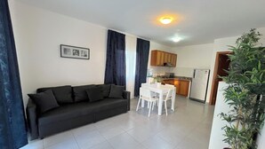Una sala de estar con un sofá oscuro, una mesa de comedor blanca y una cocina al fondo.