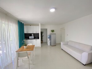 A modern living room with a white sofa, a dining table with chairs, a kitchen area with white cabinets, a refrigerator, and a potted plant. - CARIBBEAN STUDIOS playa LOS CORALES (Punta Cana)