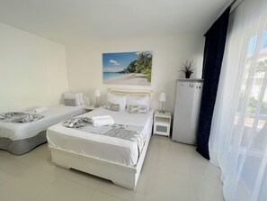 A hotel room with two single beds, a large bed, a small refrigerator, a nightstand, and a painting of a beach scene. - CARIBBEAN STUDIOS playa LOS CORALES (Punta Cana)
