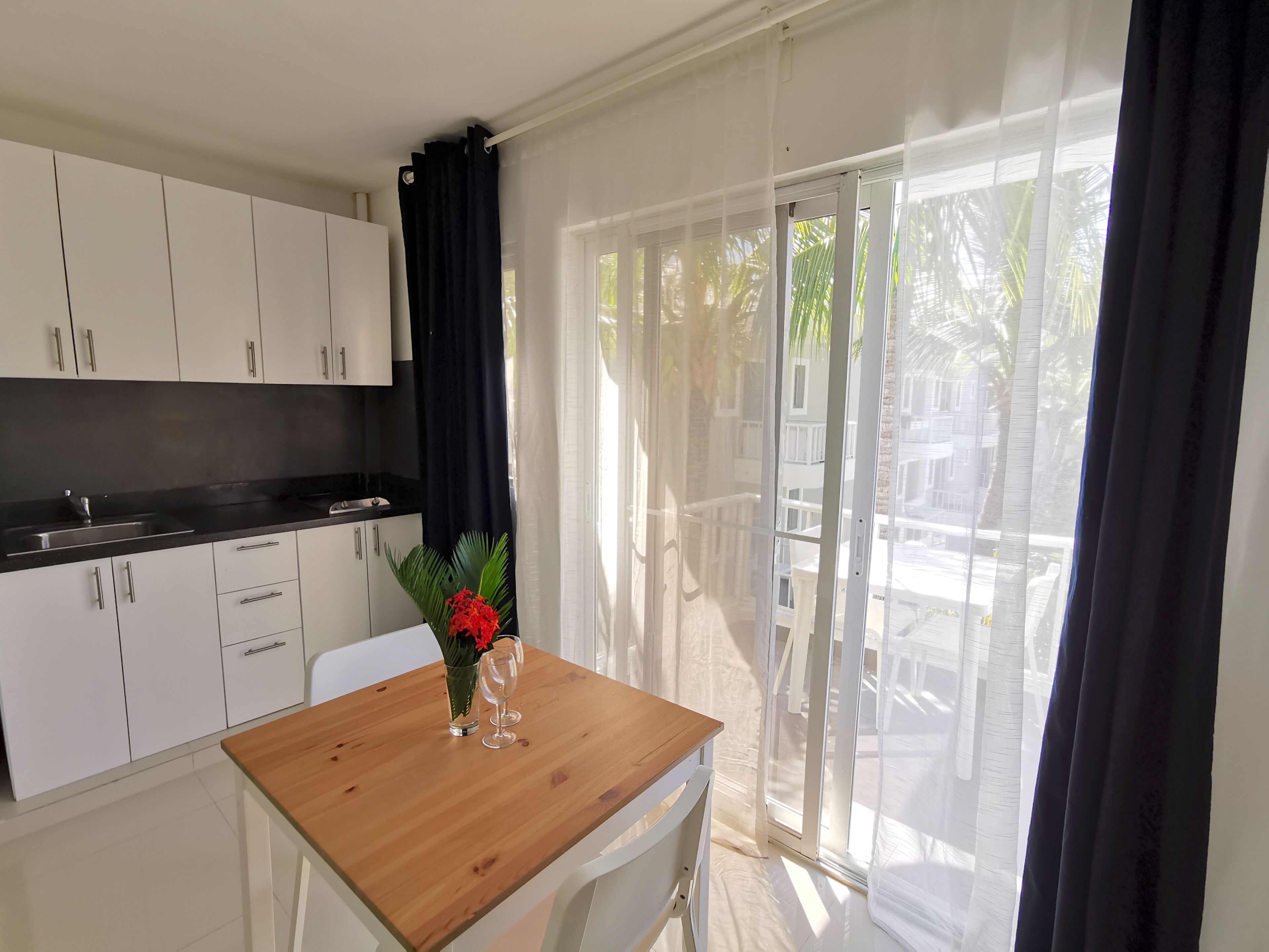 Una cocina moderna con armarios blancos y una encimera negra, una mesa de madera con un jarrón de flores rojas y un ventanal corredero que da a un área exterior.
