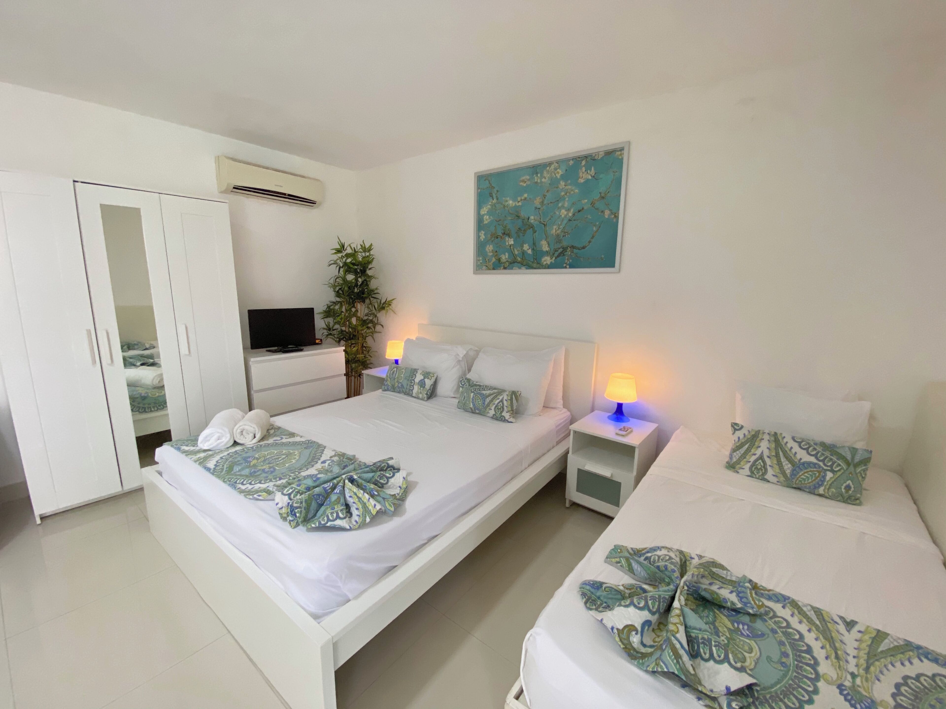 A hotel room with two beds, a TV, a plant, and a painting on the wall.