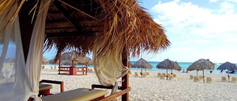 Une plage avec des parasols en chaume, des chaises longues en bois et une structure en bois dotée d’un banc.