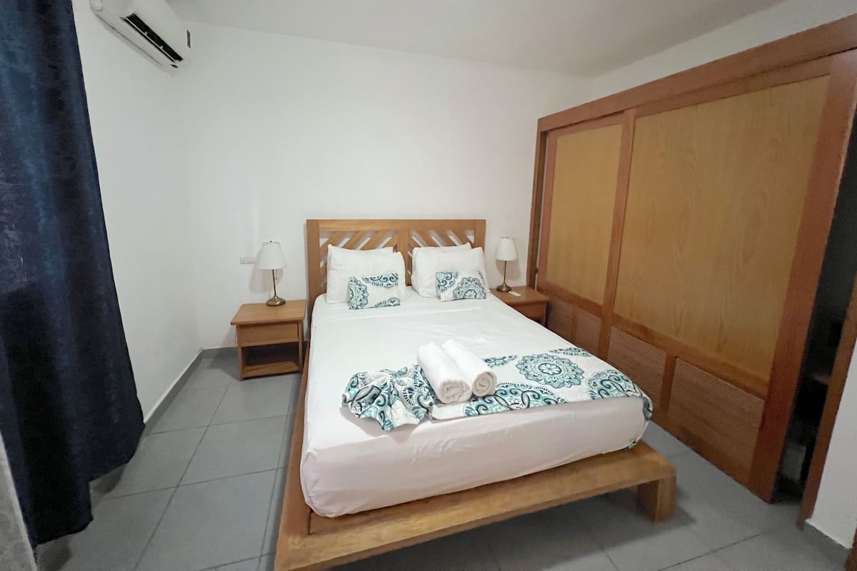 Une chambre à coucher avec un lit en bois, du linge de lit blanc, des coussins à motifs, deux lampes de chevet, une table de chevet en bois et une armoire.