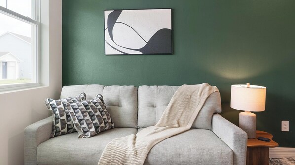 Un salon avec un canapé gris, des coussins à motifs, un plaid en tricot, une œuvre d’art abstraite accrochée au mur et une lampe de table.