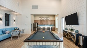 Ein modernes Wohnzimmer mit Billardtisch, einem blauen Sofa und einer Küchenzeile im Hintergrund.
