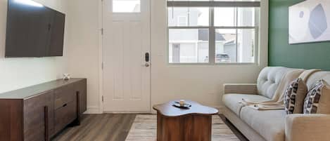 A living room with a flat-screen TV mounted on the wall, a wooden coffee table, a sofa, and a window with blinds.