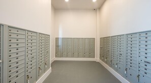 Une rangée de boîtes aux lettres métalliques dans un couloir, avec un mur blanc uni et un sol gris.
