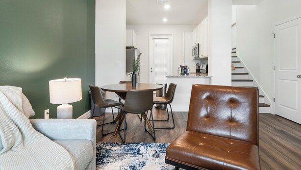 Ein Wohnzimmer mit einer Couch, einem Couchtisch und einem braunen Ledersessel. Es gibt eine Essecke mit Tisch und Stühlen, und im Hintergrund eine Küche.