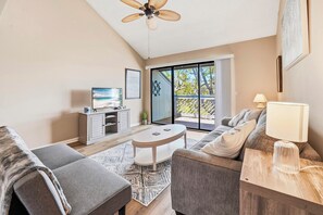 A modern living room with a sofa, a coffee table, a TV on a cabinet, and a view of the outdoors through a glass door. - Skiff F7 in Saint Simons Island, GA (Saint Simons Island)