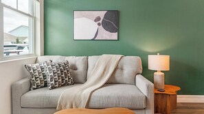 Ein Wohnzimmer mit einer Couch, Kissen, einer Decke, einer Lampe und einem gerahmten abstrakten Gemälde an der Wand.