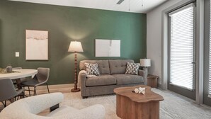 Ein modernes Wohnzimmer mit grüner Akzentwand, einem grauen Sofa mit gemusterten Kissen, einem runden Couchtisch und einem Essbereich mit weißem Tisch und grauen Stühlen.