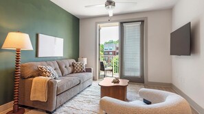 A modern living room with a sofa, a coffee table, and a chair. There is a ceiling fan, a lamp, and a TV mounted on the wall. - Landing | Modern 1BD, Gym, Yoga Studio (Charlotte)