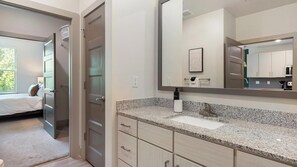 Un baño moderno con encimera de granito, un espejo grande y una zona de ducha visible.
