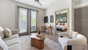 A modern living room with a sofa, a coffee table, and a TV. There is a bed in the background and a door leading to another room. - Landing | Stylish 1BD, Gym, Clubhouse (Charlotte)