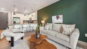 A modern living room with a sofa, armchair, and coffee table. There is a kitchen area with bar stools and a dining table. - Landing | Incredible 1BD, Gym, Yoga Studio (Charlotte)