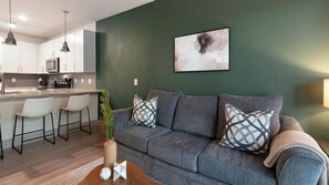 A modern living room with a grey sofa, two white chairs, a coffee table, and a green wall with a painting. - Landing | Chic 1BD, Gym, Yoga Studio (Charlotte)