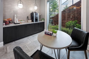 A modern kitchen with a coffee machine, a dining table with chairs, and a view of a brick wall and greenery outside. - Landing | Spectacular 1BD, Gym, Clubhouse (Charlotte)